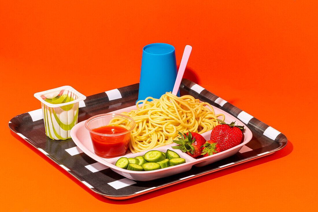 Front view of spaghetti with tomato sauce fresh cucumber slices strawberries and fruit jelly cup with empty glass for water served for school lunch