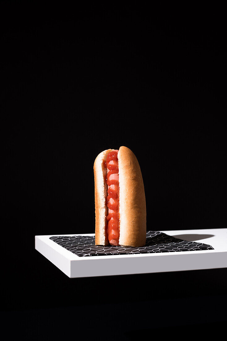 Appetizing bun with sausage and ketchup served table mat over white wooden board against black background in studio