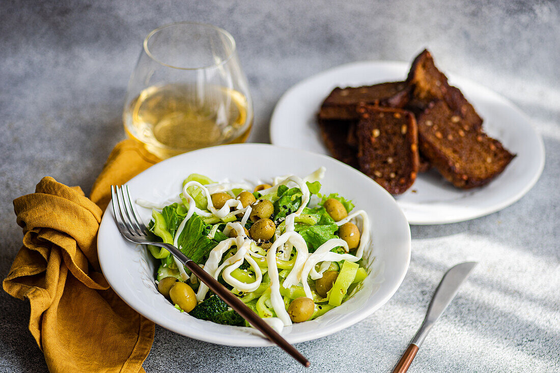 High angle of vegetable salad with green vegetables like lettuce, cucumber, olives, green bell pepper with homemade cheese and sesame seeds placed near plate of bread, glass of wine and cutlery against gray background