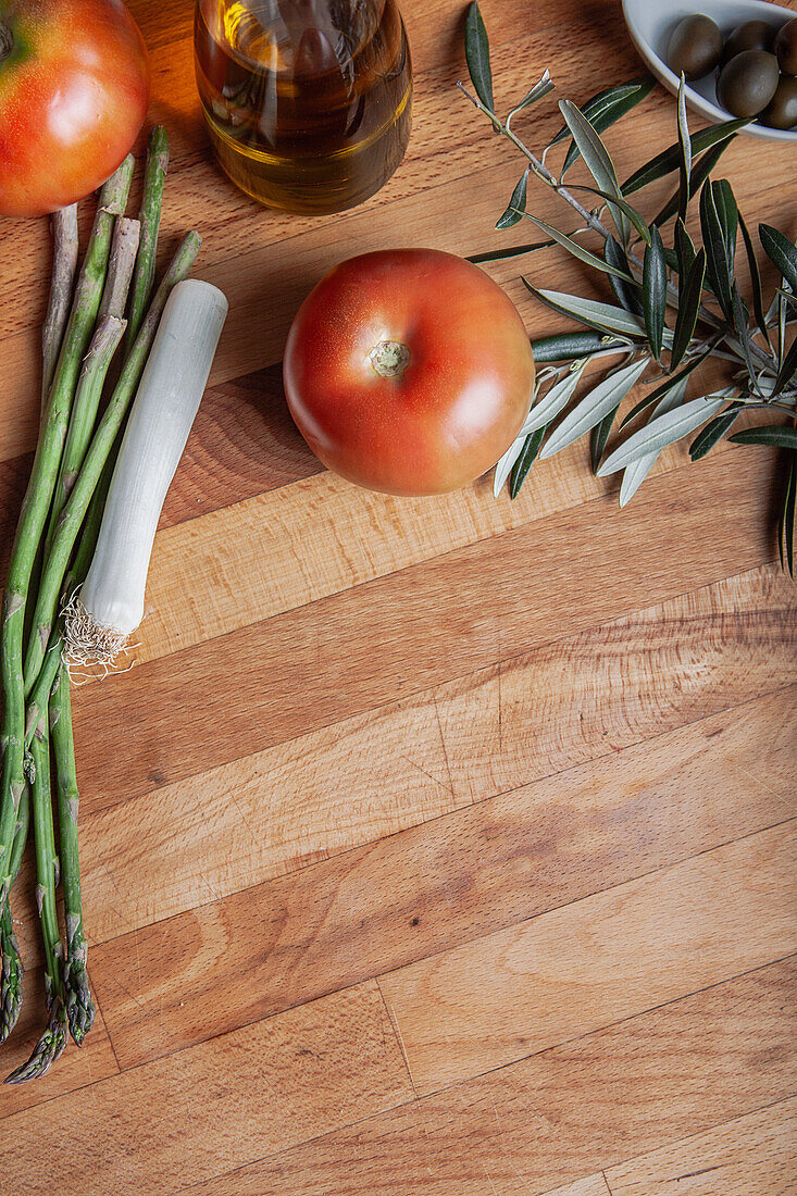 Reife Tomaten, Spargel und Lauch mit Olivenöl und Oliven auf einem rustikalen Holzschneidebrett, ideal für kulinarische Hintergründe