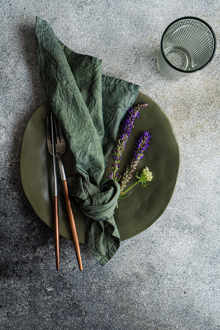 Draufsicht auf einen herbstlich gedeckten Tisch mit marmoriertem Teller, Serviette, Gabel und Messer und Lavendelblüten neben einem leeren Glas vor grauem Hintergrund