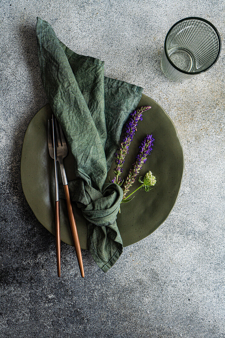 Top view of autumnal table setting with marbled plate, napkin, fork and knife and lavender flowers near empty glass against gray surface