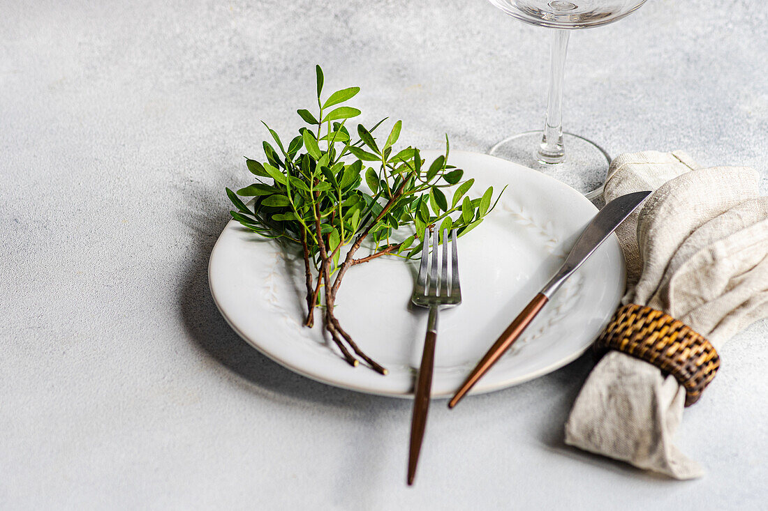 Tischdekoration mit frischer Pistazienpflanze auf einem Teller mit Besteck neben einer Serviette und einem Glas auf einer grauen Fläche im Tageslicht
