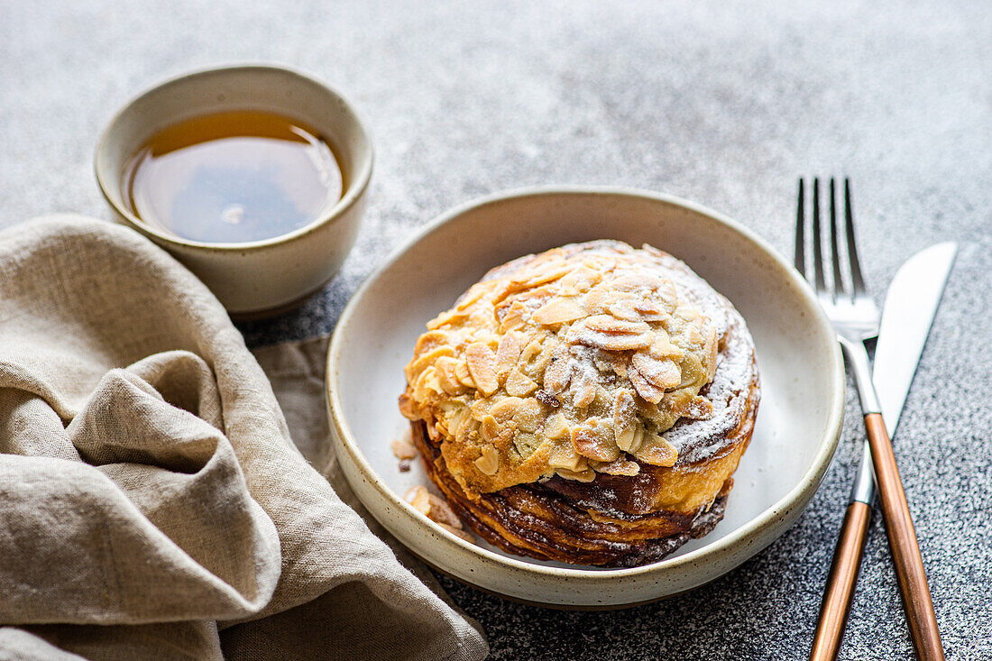 High angle of sweet almond pastry between fork and knife and napkin with cup of green tea against gray background