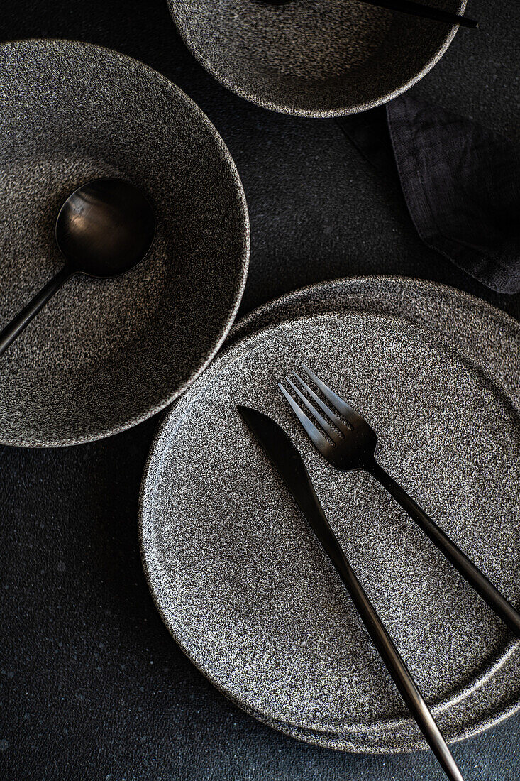 An artistic shot of a modern tableware set, including plates and utensils, arranged on a dark, textured surface.