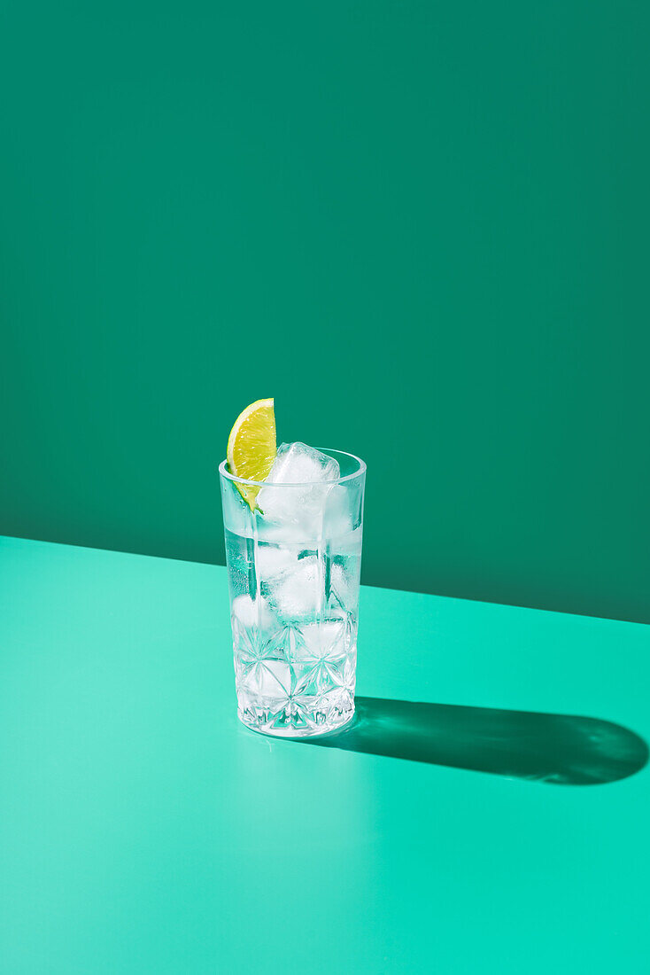 Ein mit Gin Tonic gefülltes und mit einer Limettenscheibe gekröntes Glas steht vor einem leuchtend blaugrünen Hintergrund und wirft einen scharfen Schatten