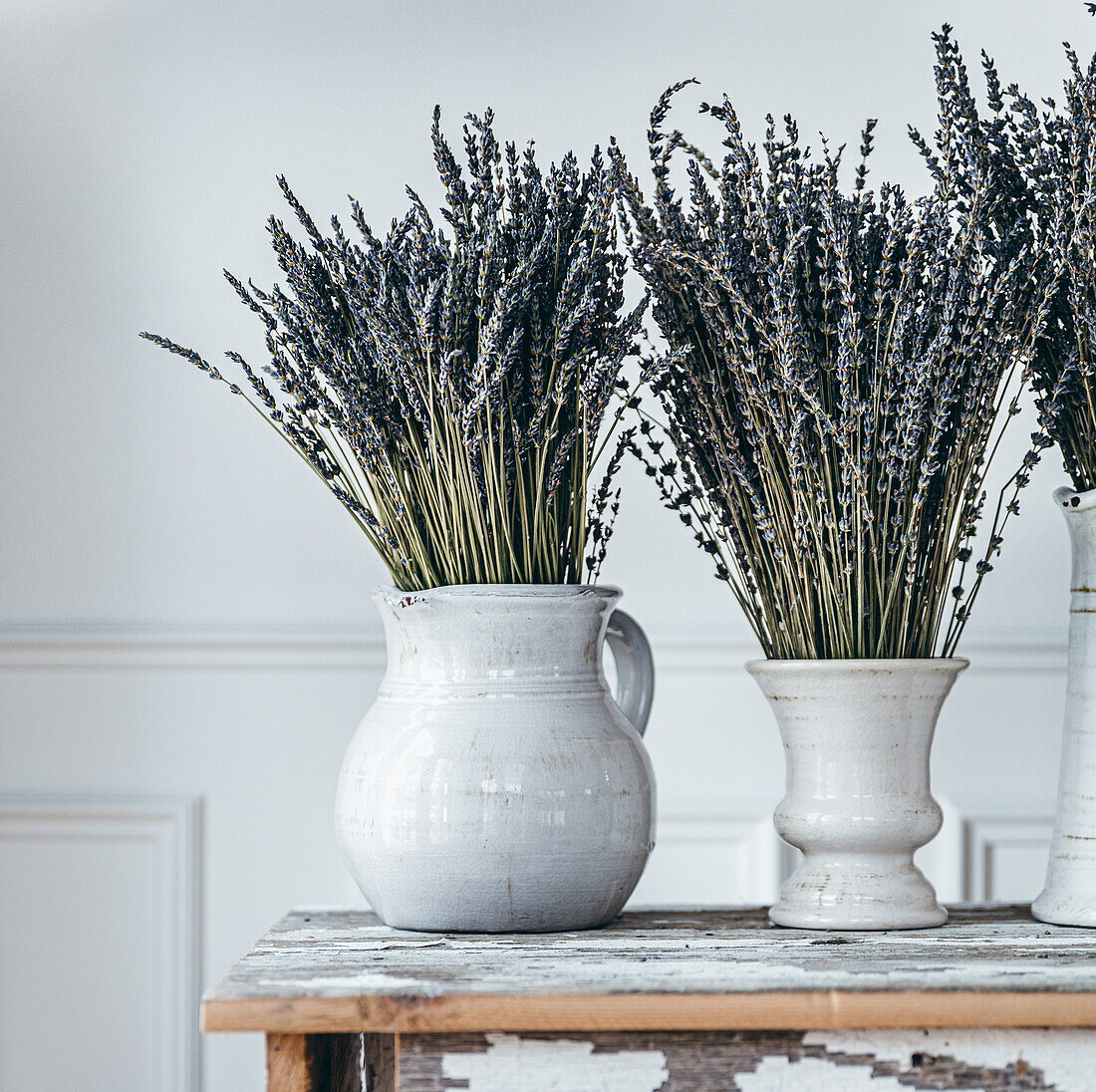 White ceramic vases with branches of blooming lavender flowers placed on wooden table against light wall