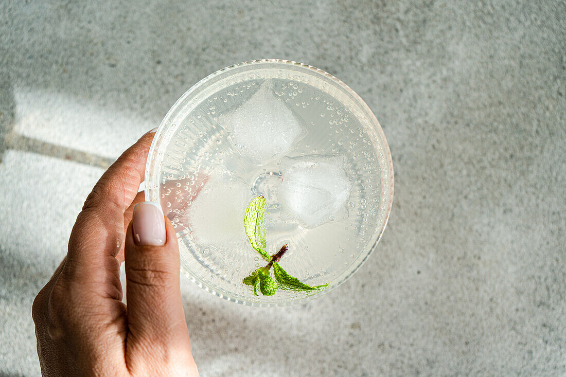 Close-up of a transparent drink with ice, lime slice, and mint in a glass cup, held by a person's hand over a grey surface