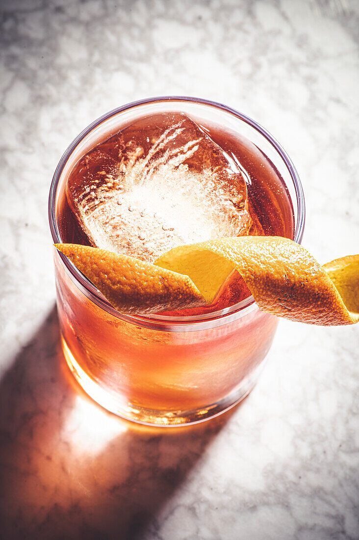 High angle of glass filled with old fashioned cocktail and ice cubes decorated with orange peel placed on marble surface