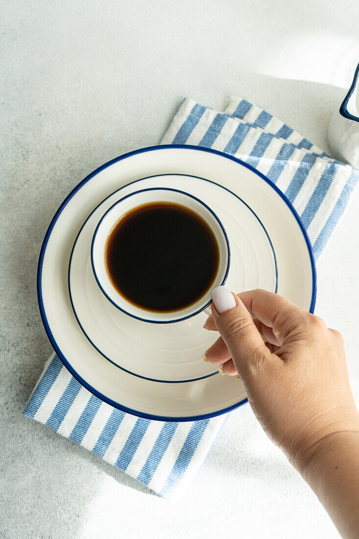 A person's hand is holding a white cup filled with drip coffee, placed on a saucer with a blue striped rim, resting on a striped napkin.