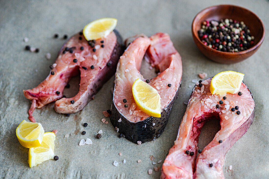 High angle of fresh trout steaks arranged on baking paper, ready to be cooked with Slices of lemon, salt and pepper near a small bowl of mixed fermented capers