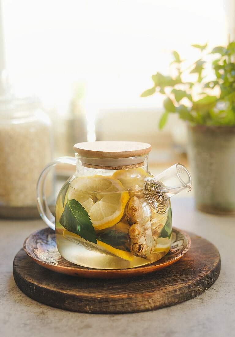 Gläserne Teekanne mit Zitrone, Minze und Ingwer auf hölzernem Schneidebrett vor Fensterhintergrund mit natürlichem Licht. Gesunder Tee mit Vitamin C in der kalten Zeit. Frontansicht