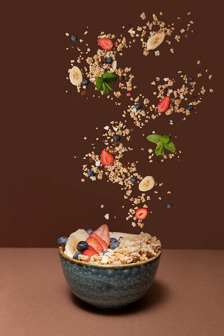 Bowl with delicious muesli and fresh berries with sliced banana splashing in air above plain brown surface against brown studio background
