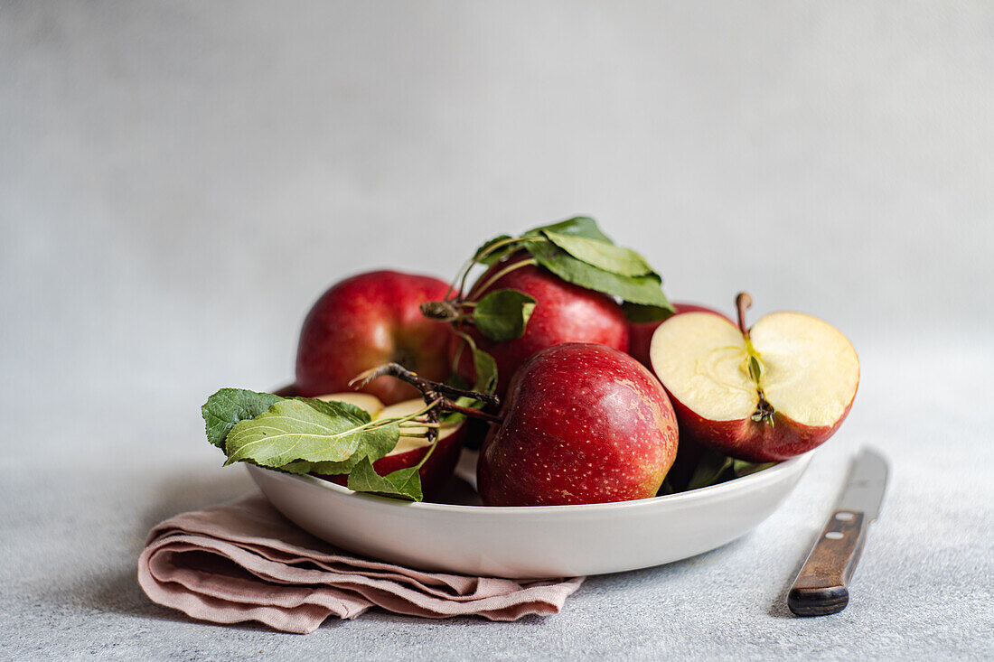 Ein Bündel reifer roter Äpfel in einem Teller mit einem Messer und einem Tuch auf einer grauen Fläche vor einem unscharfen Hintergrund