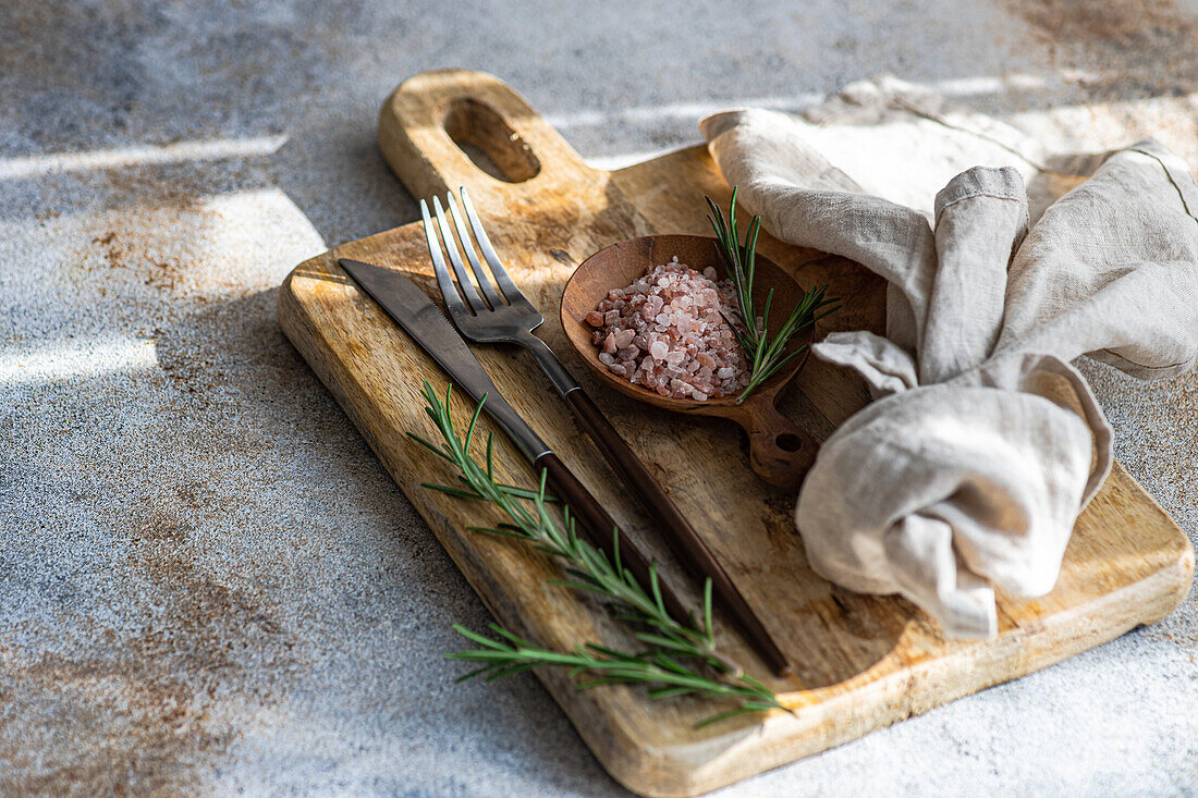 From above rustic kitchen setup on wooden cutting board with a fork, knife, a sprinkle of pink Himalayan salt, fresh rosemary sprigs, and a linen napkin, laid out on a textured concrete background