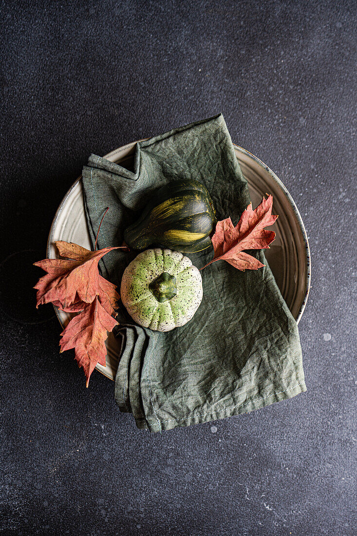 Top view of autumnal table setting with napkin, leaves and pumpkins placed on ceramic plate against dark surface