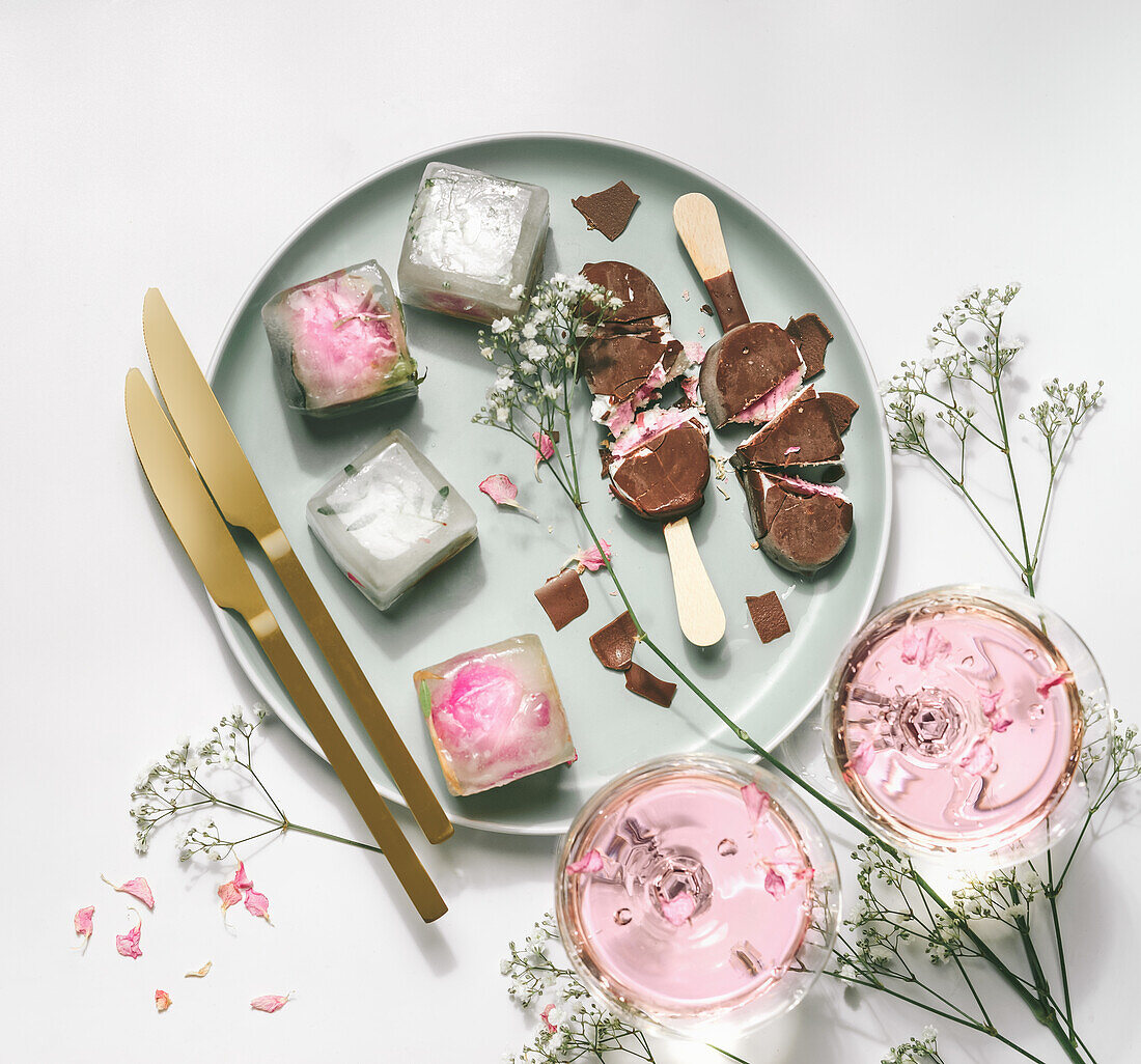 Ice cream popsicles with chocolate on plate with gypsophila, wine glasses with rose wine, ice cubes and golden knifes at white table. Summer setting with refreshing ice cream and champagne. Top view.