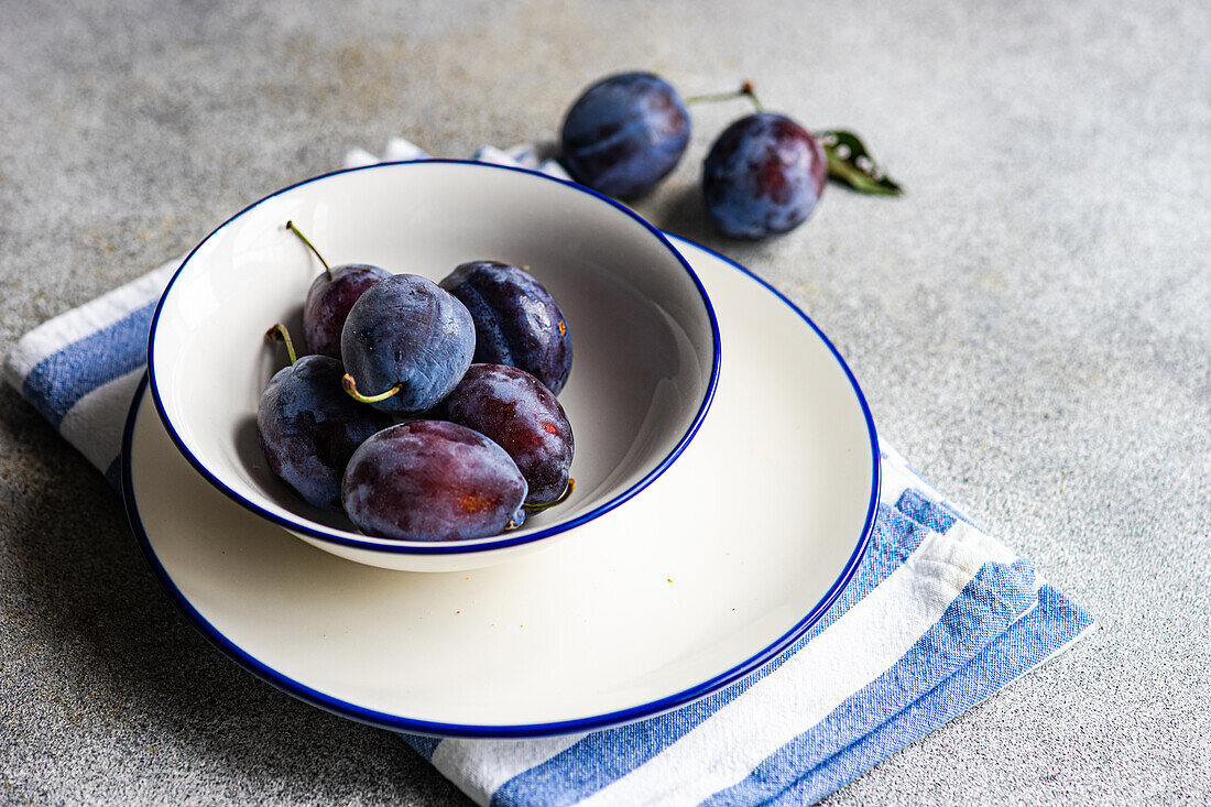 Ripe plums served in ceramic bowl on striped napkin against gray surface