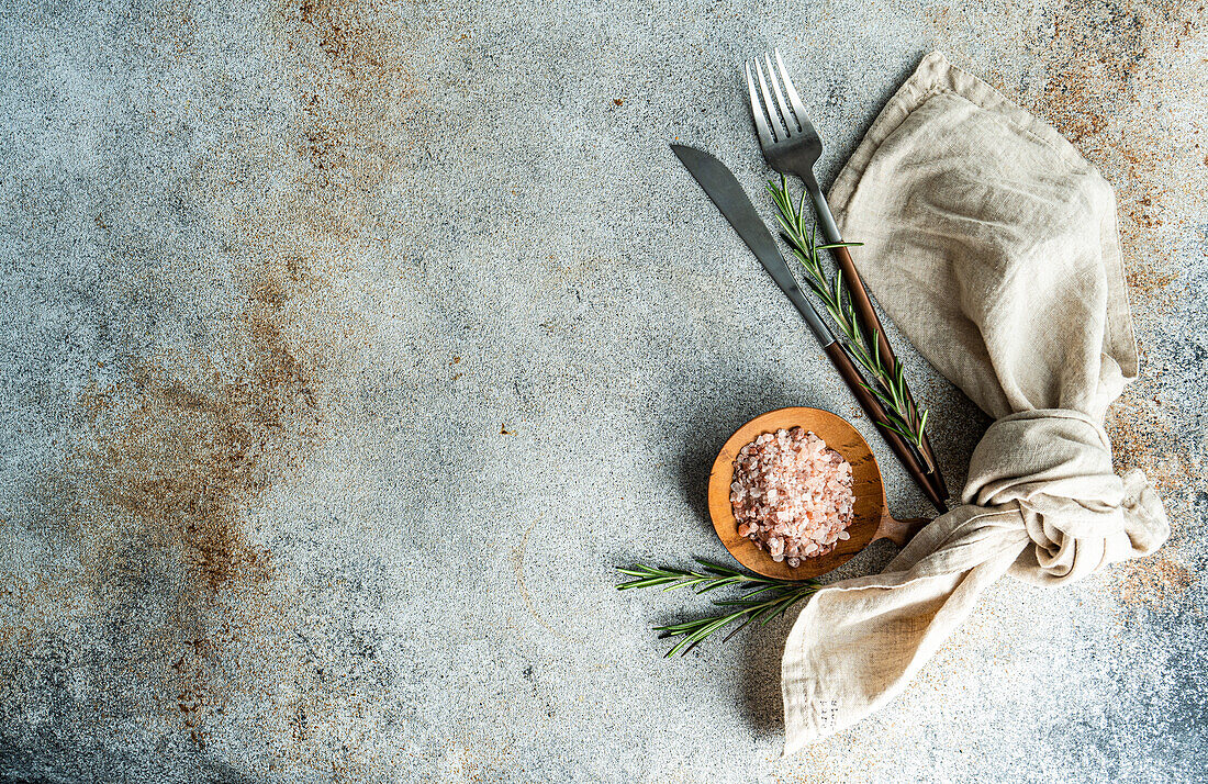 From above wooden spoon with pink Himalayan salt, surrounded by rosemary sprigs and a folded linen napkin alongside a knife and fork, all arranged neatly on a textured concrete surface