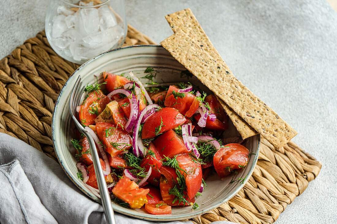 Schale mit Salat aus Tomaten, roten Zwiebeln und Dillkräutern in der Draufsicht neben einem mit Eis gefüllten Glas