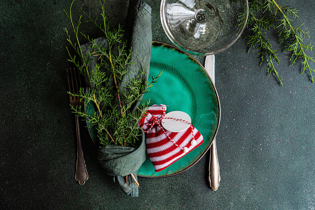 Draufsicht auf eine kleine Weihnachtstüte und in ein Taschentuch gewickelte Tannenzweige auf einem Teller, der auf einem grünen Tisch neben Glas und Besteck steht