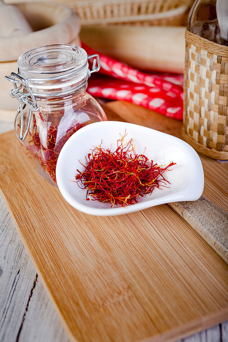 From above kitchen setup with a mortar and pestle, a checkered napkin, and saffron threads displayed on a white bowl and a glass jar, all arranged on a wooden cutting board