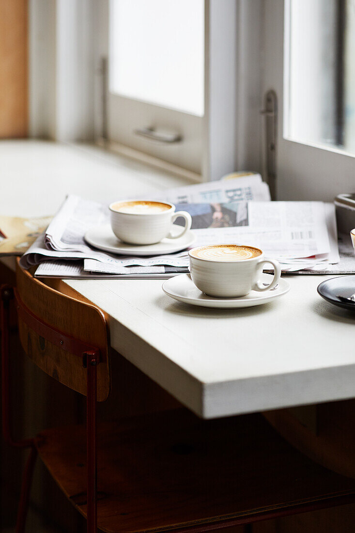 Weiße Keramiktassen mit aromatischem Cappuccino stehen auf einer Zeitung auf der Fensterbank eines Cafés