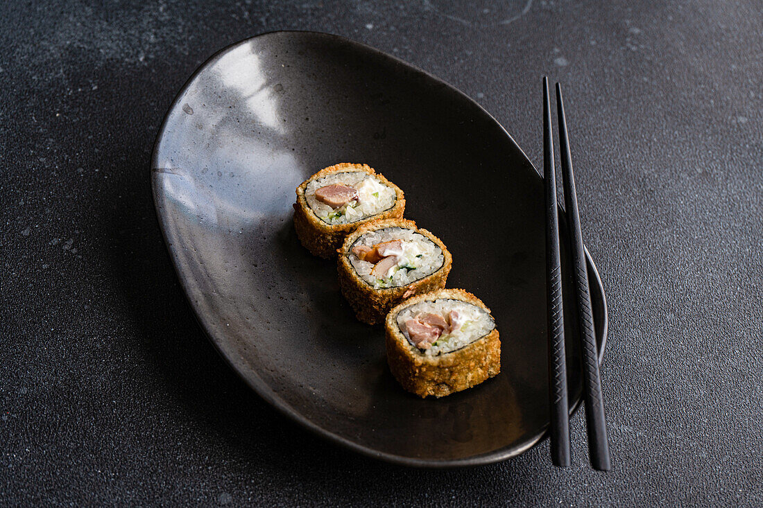 From above row of tasty fried sushi rolls with cooked rice and salmon with avocado slices on ceramic black plate on table