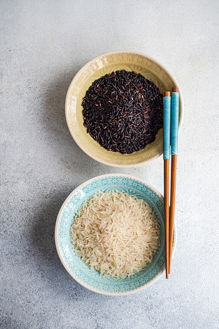Top view of Raw wild black rice and peeled white rice in decorated bowls with chopsticks against white surface