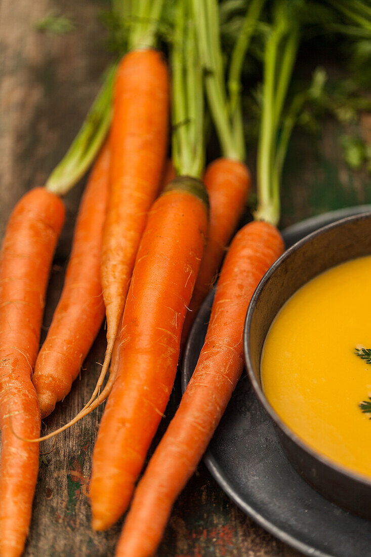 A cluster of vibrant, fresh carrots with green tops alongside a bowl of creamy carrot soup garnished with herbs on a rustic wooden surface.