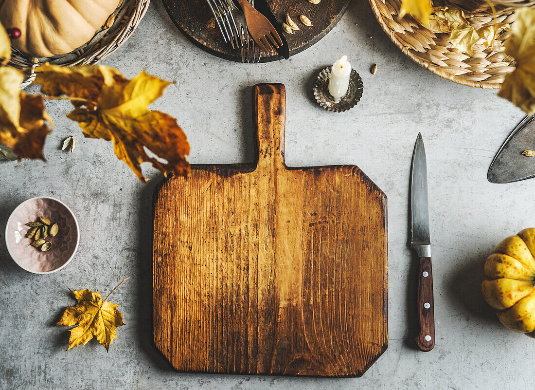 Food background with empty wooden cutting board, pumpkin, knife, autumn leaves and utensils at grey table. Cooking at home with seasonal ingredients. Top view with copy space.