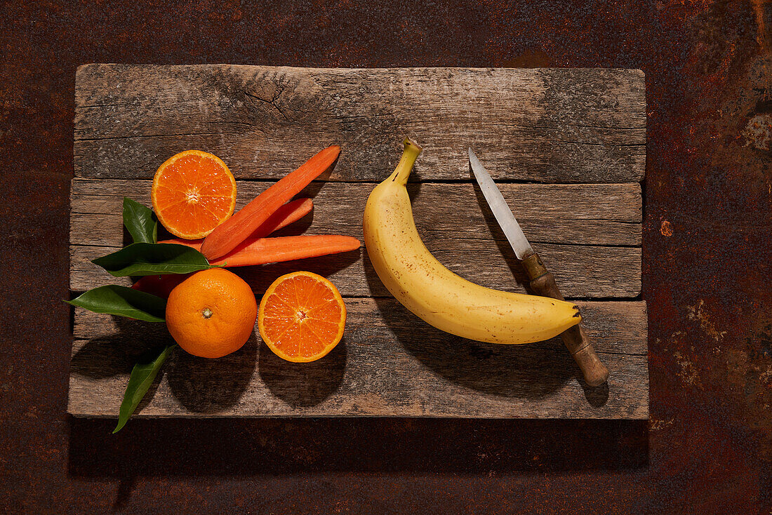 Draufsicht auf ein Arrangement aus reifen Orangen und Bananen, das neben mit einem Messer geschnittenen Karottenscheiben auf einem Holztisch steht
