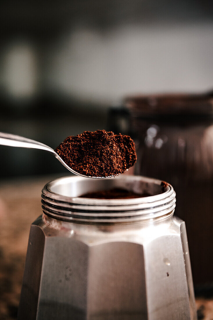 Close-up of spoon ground coffee from a jar to a stainless steel stovetop espresso maker set on a speckled countertop