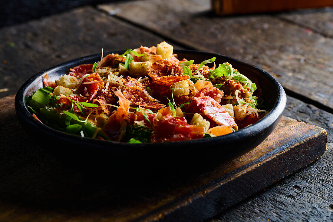 Hoher Winkel von appetitlichen Salat, bestehend aus frischem gehackten Gemüse, Kräutern und Speck in einer schwarzen Schüssel serviert