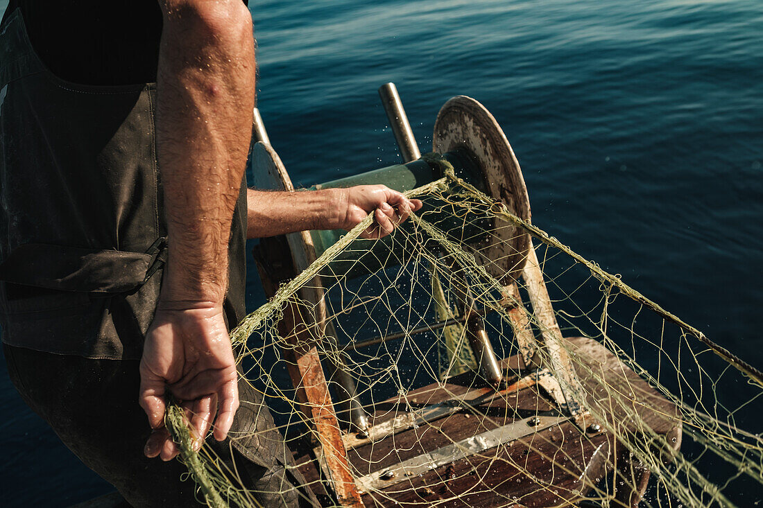 Unkenntlich gemachter männlicher Fischer in Uniform, der auf einem Schoner in Soller in der Nähe der Baleareninsel Mallorca mit einem Netz auf Fischfang geht