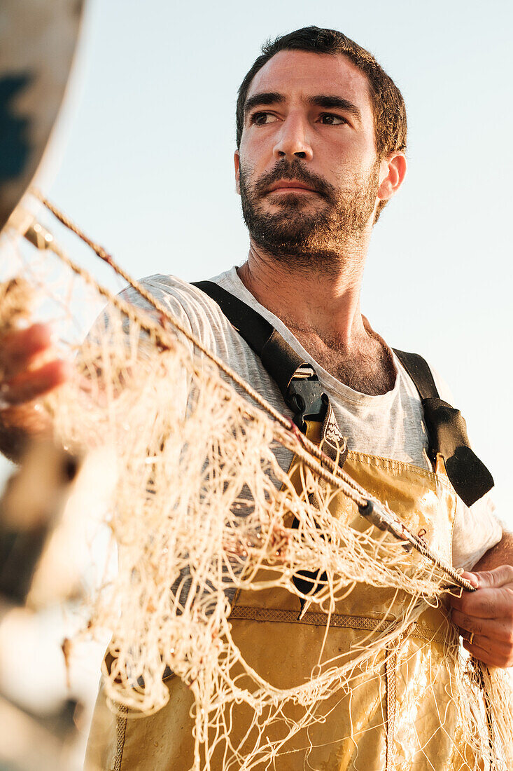 Porträt eines bärtigen männlichen Fischers in Uniform, der auf einem Schoner in Soller nahe der Baleareninsel Mallorca mit einem Netz auf Fischfang geht, von unten