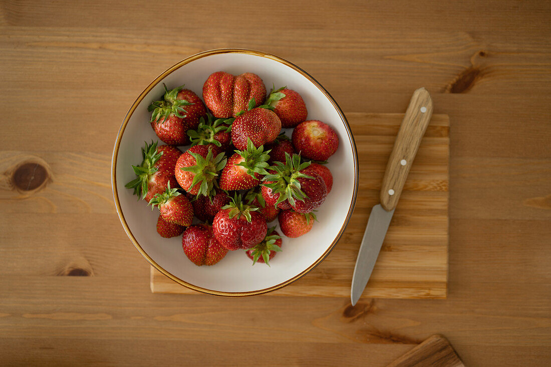 Frische Erdbeeren von oben in einer Schale und mit einem Messer auf einem Holzbrett über einem Holztisch in einem Innenraum bei Tageslicht