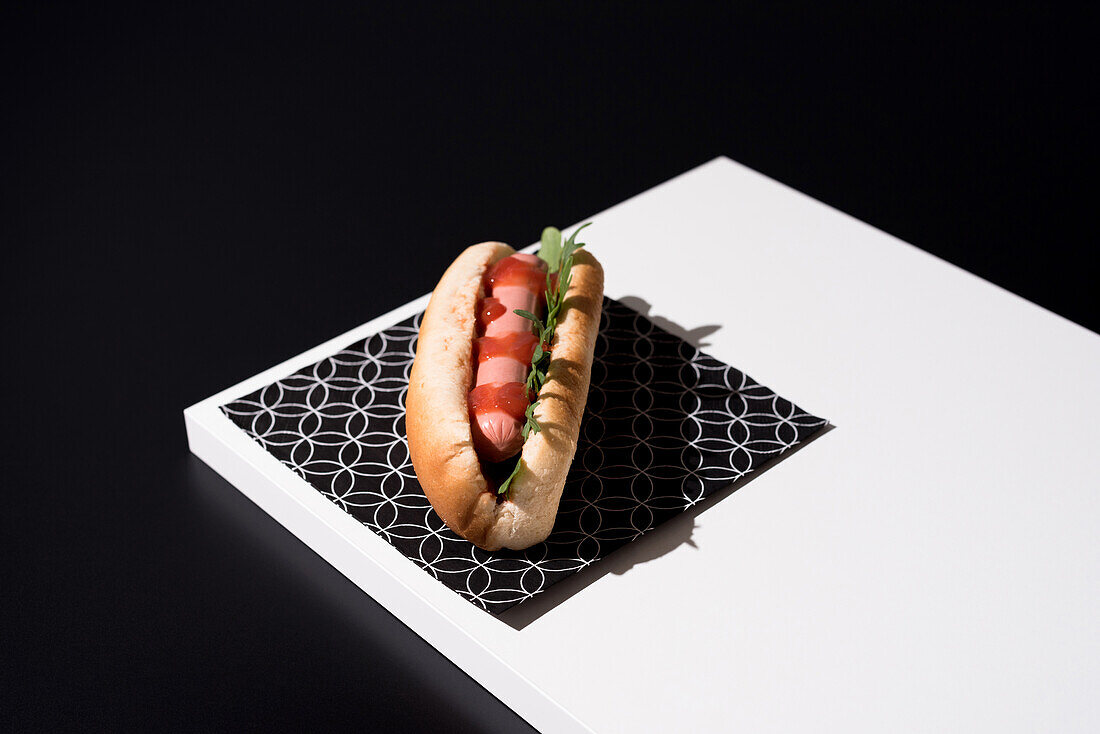 High angle of appetizing bun with sausage and ketchup with greens served table mat over white wooden board against black background in studio