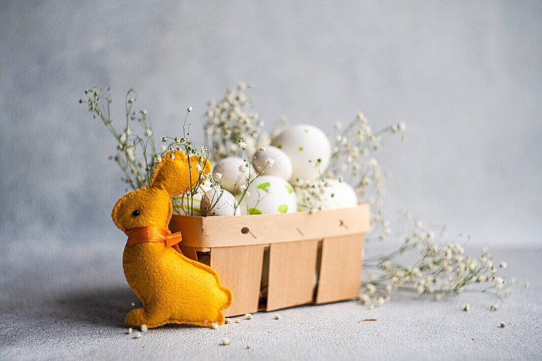 A plush yellow Easter bunny gazes at a basket filled with decorated white eggs and sprigs of baby's breath on a textured grey background