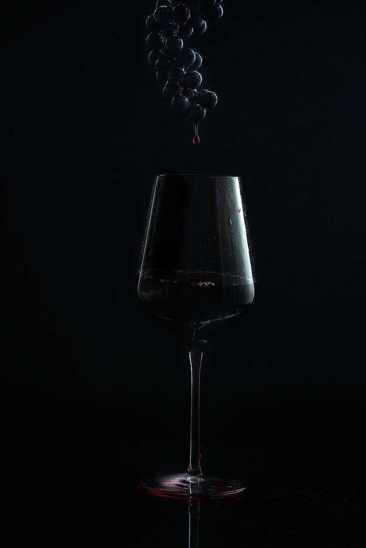 A moody shot capturing a trickle of wine descending into a glass with a dark grape bunch hovering above against a black background