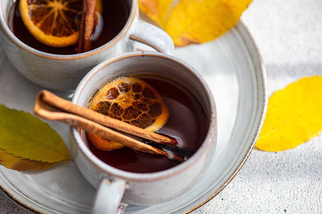 focused of two cups of aromatic spiced tea with cinnamon sticks, anise, and dried orange slices surrounded by radiant yellow autumn leaves on a textured gray background