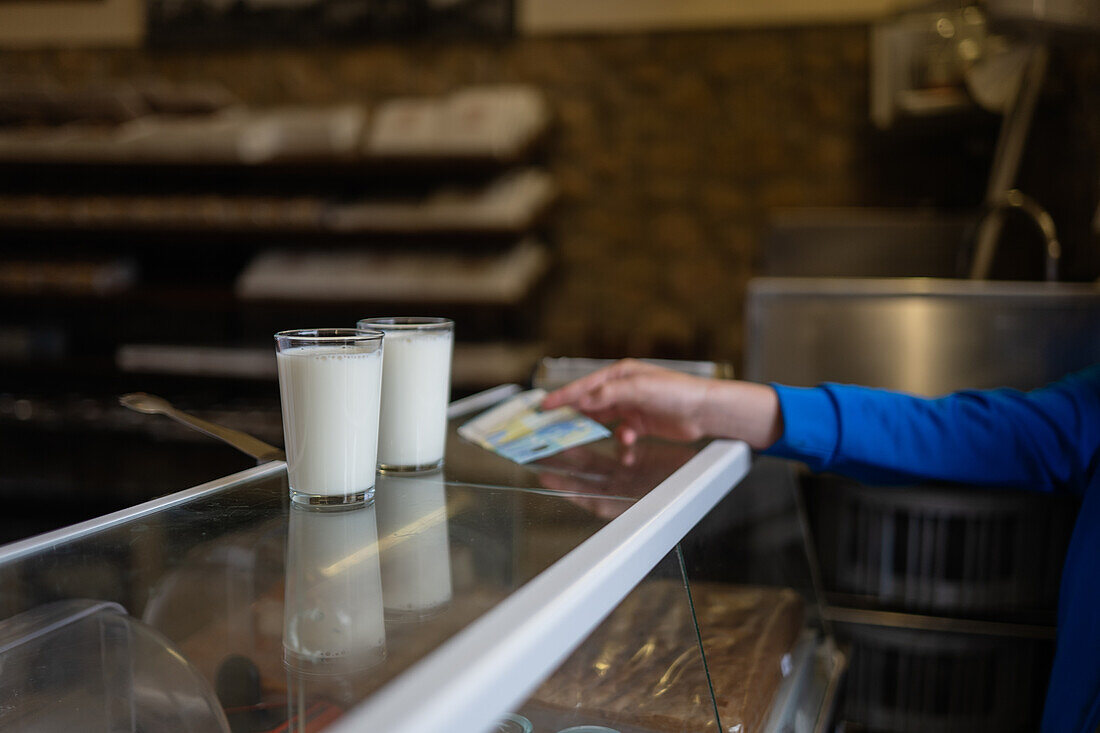 Close-up of two glasses of milk on a counter with a anonymous person hand in the background in a bakery setting