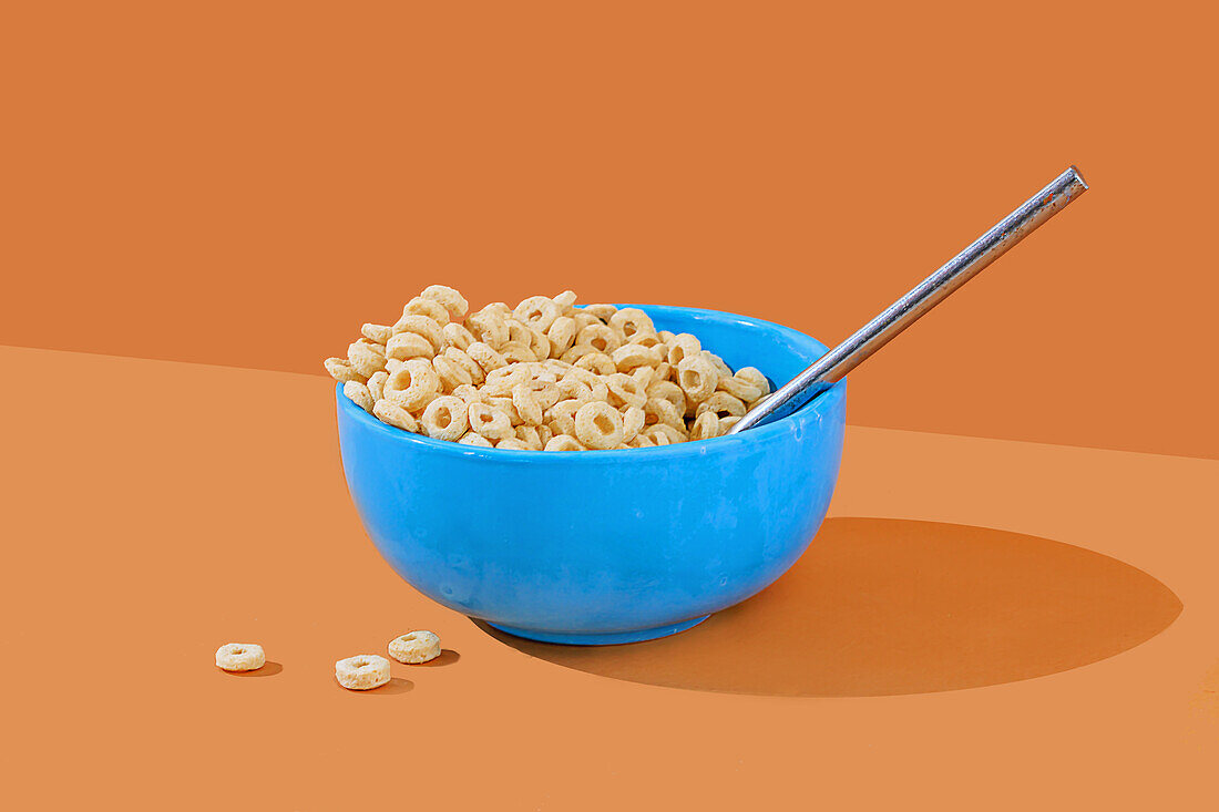 A blue bowl filled with loop-shaped cereal and milk, accompanied by a spoon, set against a vibrant orange backdrop.