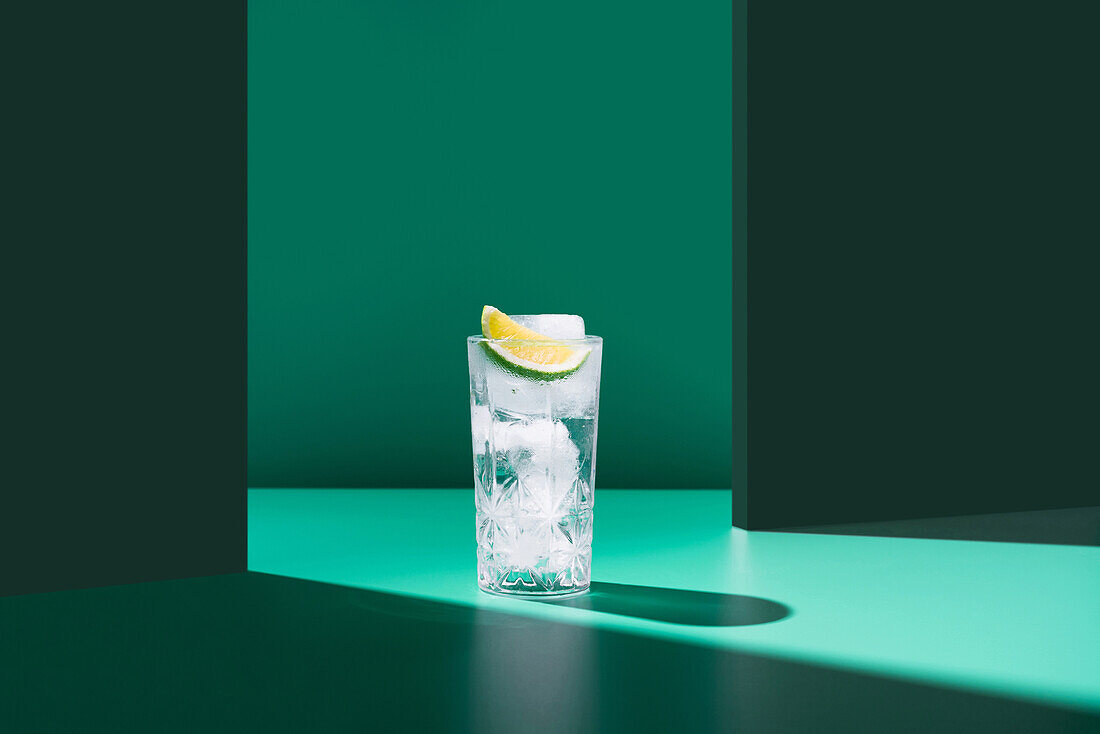 A refreshing glass of gin tonic topped with a lime slice is set against a stark green backdrop, offering a minimalist aesthetic.
