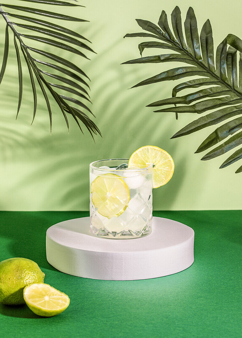 Sparkling transparent crystal glass with ice and slices of lime in cocktail drink on coaster on green surface table near leaves