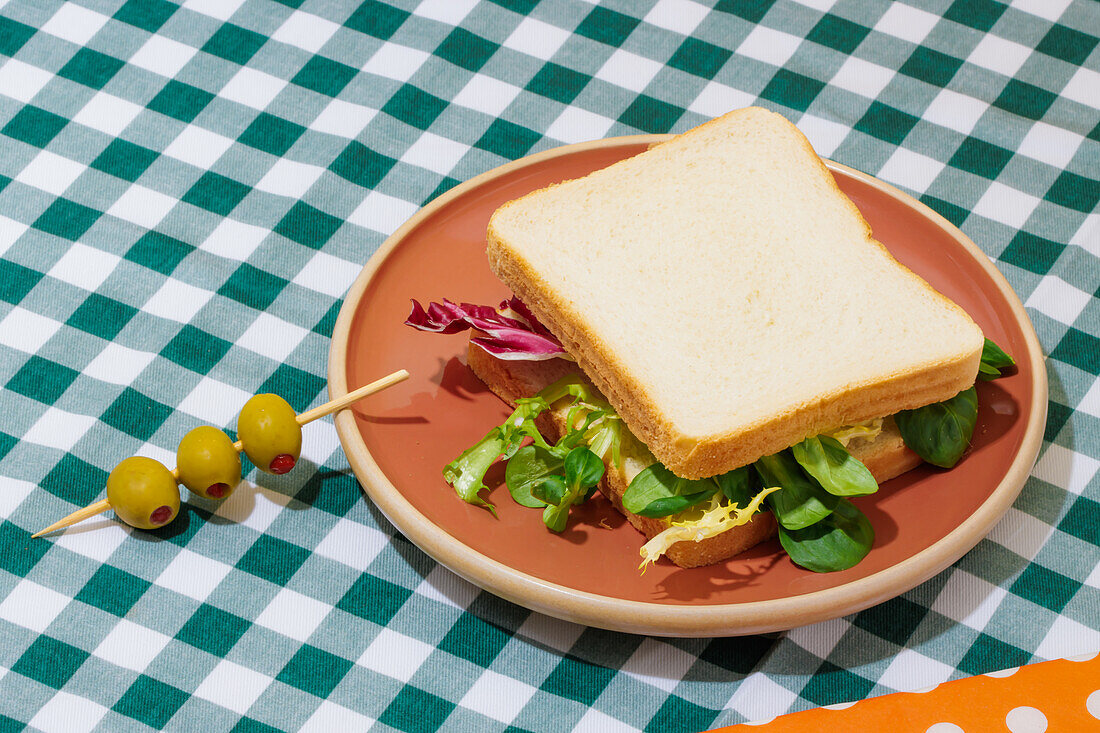 Appetizing vegetarian sandwich with fresh lettuce on plate served with olives on skewer on table with checkered tablecloth