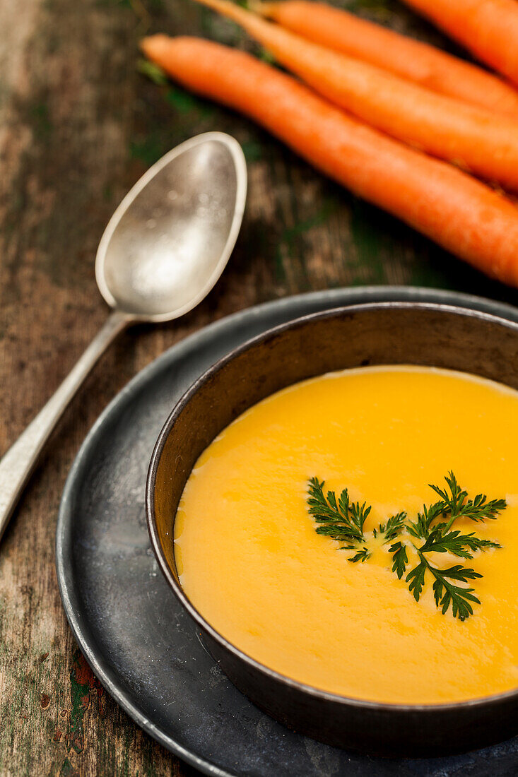 A bowl of creamy carrot soup garnished with parsley, fresh carrots, and a vintage spoon on a wooden table.
