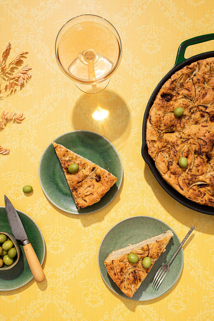 Draufsicht auf flaches, gesäuertes, im Ofen gebackenes italienisches Focaccia-Brot in einer Pfanne, serviert mit frischen grünen Oliven und Honig in Schalen auf gelber Hintergrundfläche