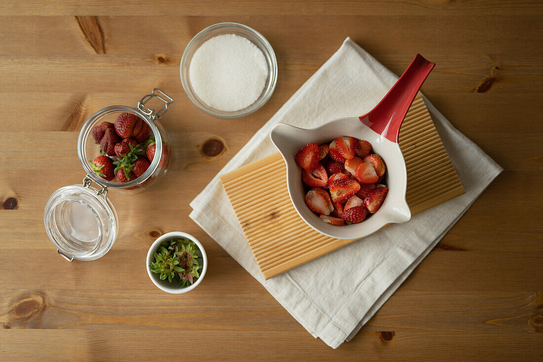 Draufsicht auf köstliche, frisch geschnittene Erdbeeren und Zucker in einer Glas- und Keramikschale auf einem Holztisch mit Stoff während der Marmeladenzubereitung in einem Raum