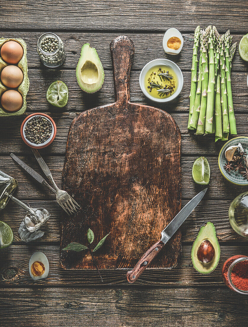 Food background frame with various healthy ingredients: green asparagus, avocado halves, spring onion, cooked eggs halves , herbs and spices around wooden cutting board with knife and forks. Top view